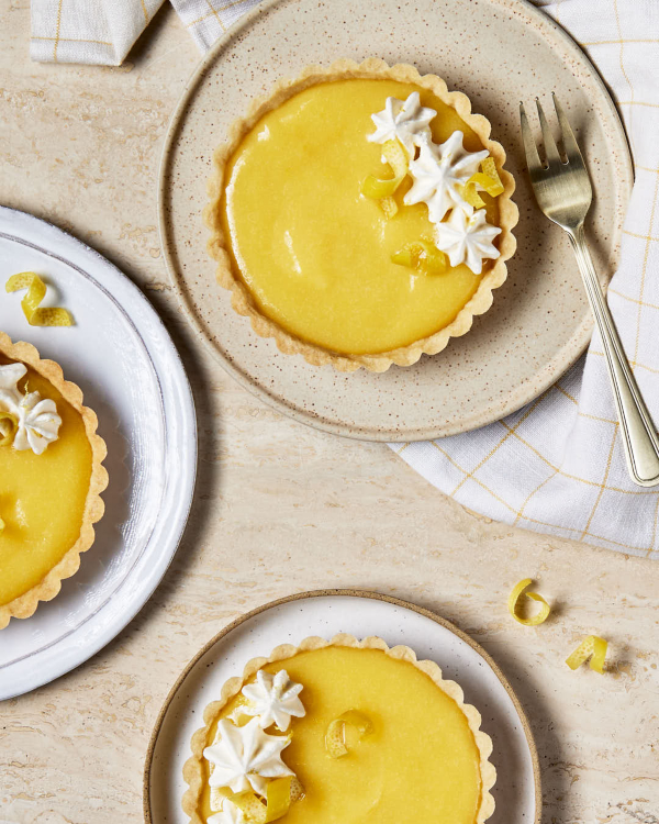 Three lemon curd tarts garnished with whipped cream and lemon zest shown on a marble counter.
