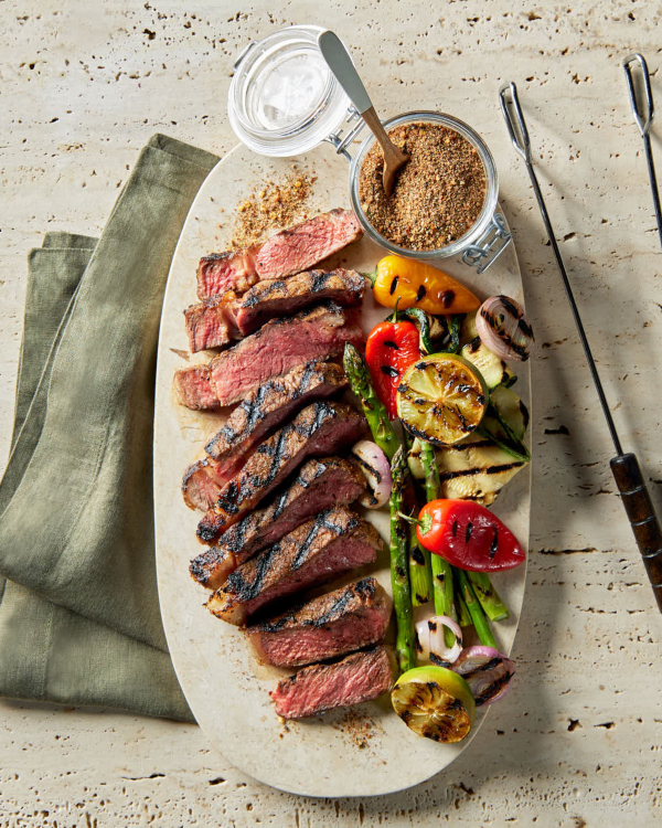 A platter of grilled and sliced steak and grilled vegetables with a lidded glass jar of sweet Montreal steak seasoning shown with barbecue tongs on a limestone counter.