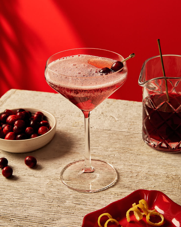 A stemmed glass filled with a carbonated cranberry cocktail, garnished with a skewer of cranberries and lemon zest, shown with a glass pitcher of cranberry cocktail, a dish of cranberries, and lemon zest on a red plate.