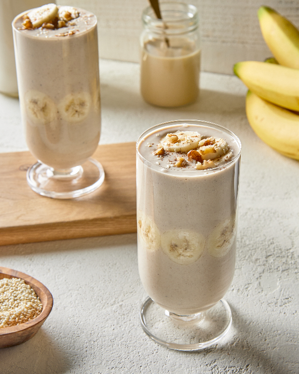 Two tall glasses of banana oat tahini smoothie, one on a wooden cutting board, shown with a bowl of sesame seeds, a jar of oat milk beverage, and a bunch of bananas.