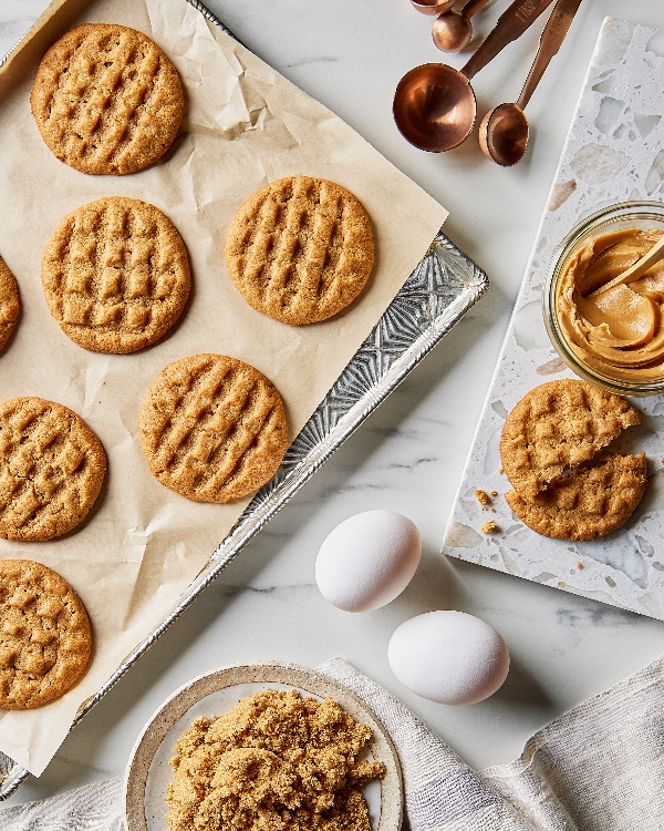 Eight homemade peanut butter cookies with golden-brown surfaces and traditional fork marks, accompanied by a jar of peanut butter, measuring spoons, and two whole eggs on a kitchen countertop.