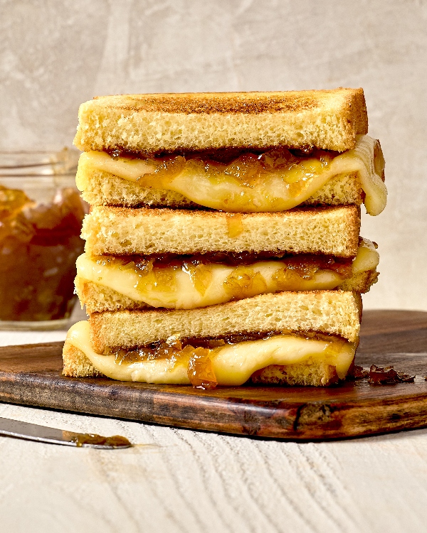 A stack of Onion Jam Grilled Cheese Sandwich halves, placed on a wooden board. The sandwiches have golden-brown toasted bread with melted cheese and caramelized onion jam oozing out. In the background, a jar of onion jam and a knife with jam are partially visible