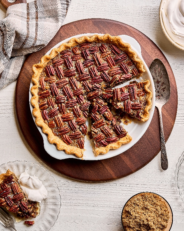 A pecan pie in a baking dish, shown with a slice being served on a pie lifter by a person in an apron, on a white wood table with a bowl of pecans