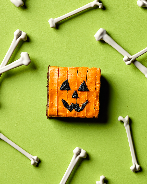 A chocolate brownie decorated with icing and to look like a jack o’lantern, shown on a green background with bones scattered around.