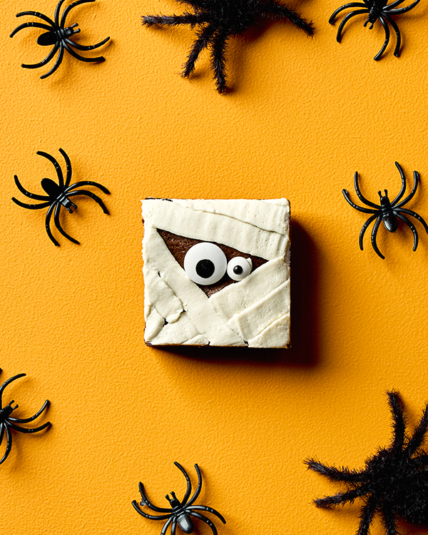 A chocolate brownie decorated with icing and candy eyes to look like a mummy, shown on a yellow background with toy spiders scattered around.