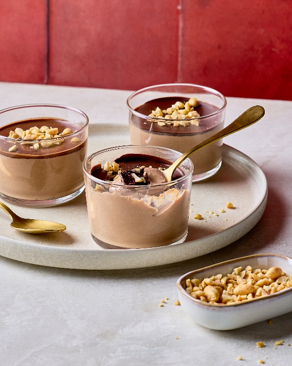 Three servings of chocolate peanut butter pudding in glass jars, topped with a glossy chocolate layer and chopped peanuts. One jar is partially eaten, showing the creamy interior, with a golden spoon resting inside. The jars are placed on a neutral round tray, accompanied by a small dish of chopped peanuts, set against a red background.