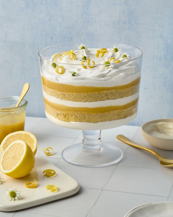 A glass trifle dish filled with a layered lemon elderflower trifle, showcasing alternating layers of sponge cake, lemon curd, and whipped cream. The top is garnished with lemon zest curls and small white flowers. Surrounding the trifle are fresh lemons, a jar of lemon curd, and a gold spoon, set against a soft blue background.