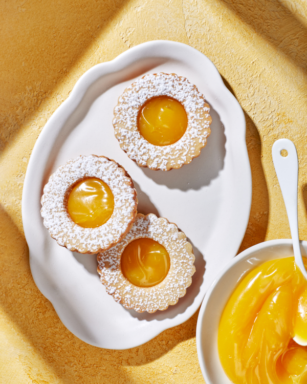 Citrus curd sandwich cookies topped with icing sugar on a white platter on a yellow surface, shown with a bowl of citrus curd