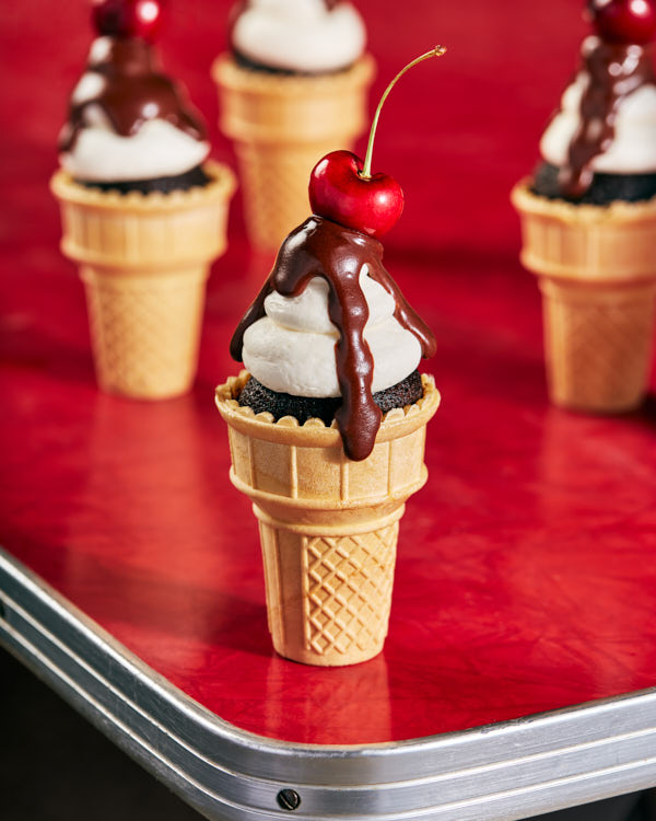 Chocolate cupcakes in classic ice cream cones decorated with icing, chocolate ganache, and topped with a cherry shown on a vintage diner tabl