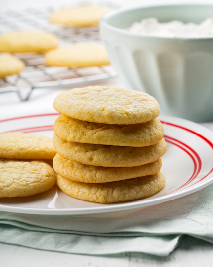 Lemon sugar cookies stacked on plate with more cookies cooling in the background