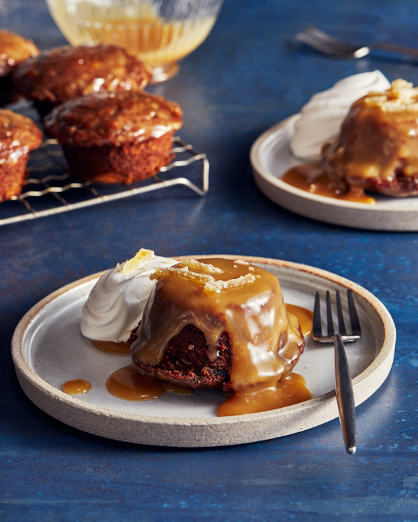 Two sticky toffee puddings on plates served with toffee sauce and whipped cream with more puddings on a cooling rack in the background.