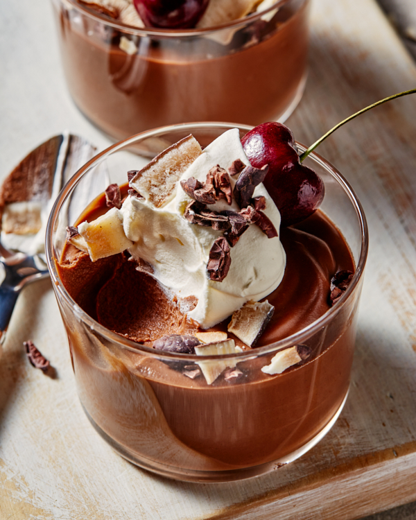 Two vegan chocolate puddings topped with whipped cream, coconut flakes, cacao nibs, and cherries