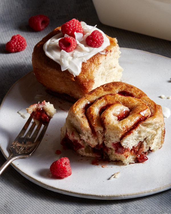 Two vegan raspberry cinnamon rolls on a plate, one with coconut glaze and raspberries, one with a forkful missing