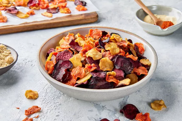 A stoneware bowl of mixed air fryer root vegetable chips shown with a bowl of Golden Yellow Sugar, a bowl of spices, and chips drying on a cutting board.