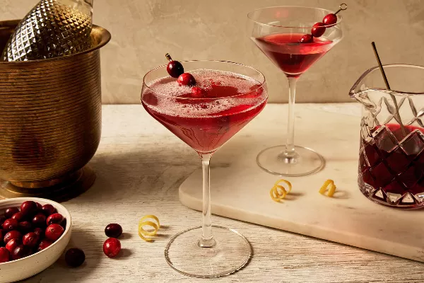 A stemmed glass filled with a carbonated cranberry cocktail, garnished with a skewer of cranberries and lemon zest, shown with a glass pitcher of cranberry cocktail, a stemmed glass of flat cranberry cocktail, lemon zest, and a bottle of sparkling wine in a brass ice bucket