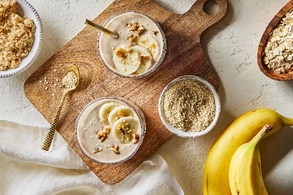  Top view of two tall glasses of banana oat tahini smoothie shown on a wooden cutting board with bowls of golden yellow sugar, sesame seeds, and dry oats with two unpeeled bananas.