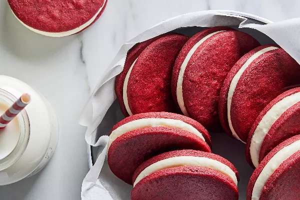 Red velvet sandwich cookies shown in a parchment-lined bowl beside a glass jar of milk with a paper straw.
