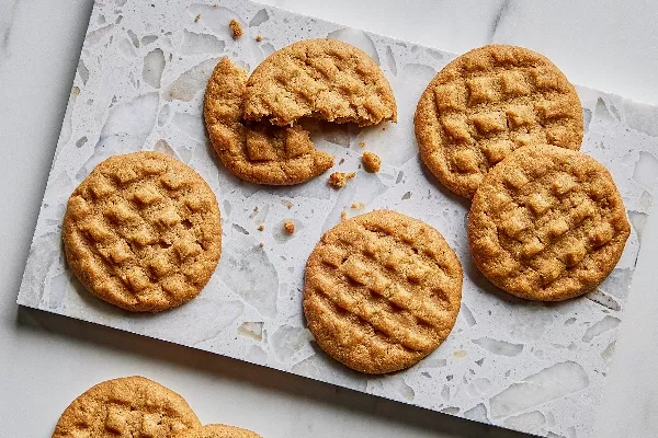 Seven golden-brown peanut butter cookies with distinctive fork press patterns displayed on parchment paper over a marble surface, one partially eaten to reveal a soft, chewy texture.