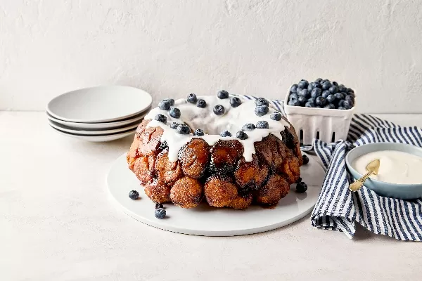 Blueberry monkey bread on a white plate with dripping white icing and fresh blueberries on top, next to a blue and white striped napkin, blue bowl with icing, and a carton of blueberries, on a light-coloured surface.