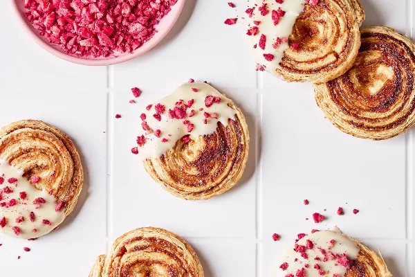 Overhead view of raspberry puff pastry cookies with white chocolate icing and freeze-dried raspberry pieces on a white tiled countertop, shown with a bowl of crushed raspberries.