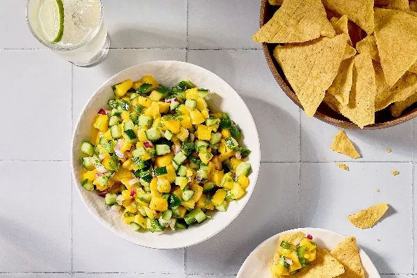 A top-down image of a bowl of mango cucumber salsa on a white tile surface. The salsa also features red onion and cilantro, and a bowl of tortilla chips is placed nearby, with some chips scattered around. A glass of lime-infused water is also visible.