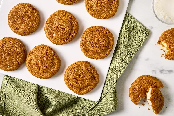 Top view of Pumpkin Spice Cream Cheese Stuffed Cookies arranged on a white tray with a green cloth underneath. The cookies have a golden-brown color and slightly cracked tops, revealing a creamy filling inside. One cookie is broken open, showing the cream cheese filling.