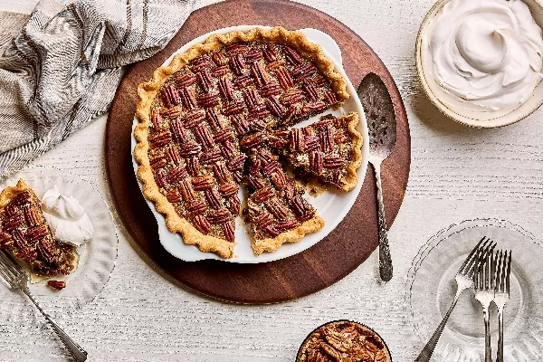 A pecan pie in a baking dish, shown with three slices cut, with one served on a plate with whipped cream, on a white wood table with a bowl of whipped cream and a bowl of pecans.