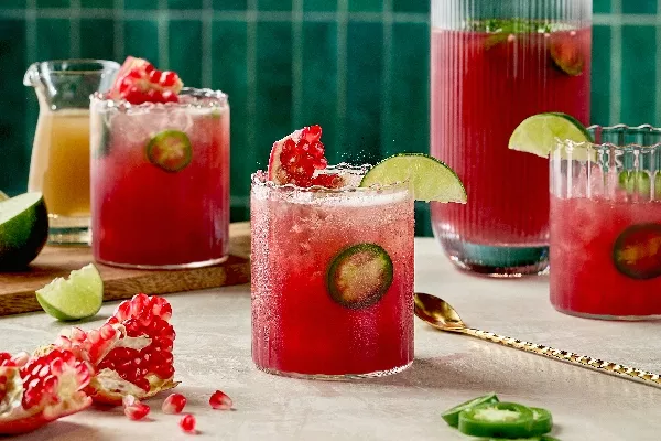 Three glasses of Spicy Pomegranate Ginger Fizz, featuring a vibrant red hue with slices of fresh jalapeño. The drinks are garnished with lime wedges and pomegranate seeds. A pitcher filled with more of the drink stands in the background, alongside lime wedges, pomegranate pieces, and a small pitcher of ginger syrup. The scene is set against an emerald green-tiled backdrop.