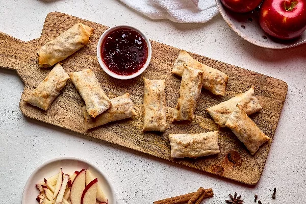 Crispy apple spring rolls arranged on a wooden serving board with a small bowl of red dipping sauce in the center. Fresh red apples and sliced apple sticks are displayed nearby, along with cinnamon sticks, star anise, and cloves