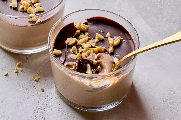  Close-up of creamy chocolate peanut butter pudding in a glass jar, topped with a glossy chocolate layer and chopped peanuts. A golden spoon rests inside, revealing the smooth and indulgent texture beneath the chocolate shell. Crumbled peanuts are scattered on the light gray surface.