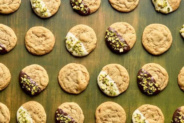 Chocolate chipless cookies arranged in a grid pattern on a green wooden surface. Some cookies are plain, while others are half-dipped in white or dark chocolate and sprinkled with chopped pistachios. 