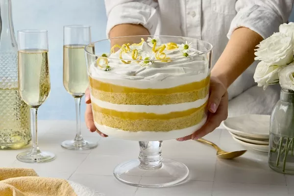 A person’s hands hold a glass trifle dish filled with a layered lemon elderflower trifle featuring alternating layers of sponge cake, lemon curd, and whipped cream. The trifle is topped with lemon zest curls and delicate white flowers. In the background are champagne glasses, a bottle, and white roses in a glass vase.