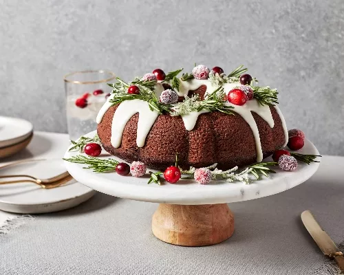 A glazed gingerbread bundt cake on a cake stand, decorated with rosemary and candied cranberries