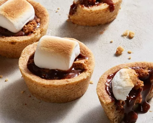 Four s’mores cookie cups with toasted marshmallows, one cut in half and leaking chocolate