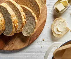 Un pain de levain sucré coupé en tranches sur une planche à découper en bois, avec une tranche beurrée, présenté avec un plat de beurre et un couteau à beurre.