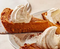 Removing a slice of pumpkin pie with whipped cream and ground cinnamon from a white pie plate with a pie lifter