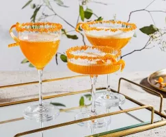 Three glasses of sparkling spiced ginger turmeric cocktail garnished with orange peel and rimmed with sugar on a mirrored tray