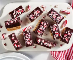 Golden cookie bars topped with chocolate ganache and peppermint stick pieces on a white tray with a red and white striped cloth