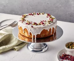 Persian Love Cake with icing and rose petals on a silver cake stand, shown with plates, forks, and napkins