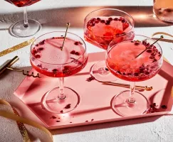 Three pomegranate-rose sparkling cocktails in stemmed glasses on a pink tray, shown with gold swizzles and gold ribbons