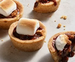 Four s’mores cookie cups with toasted marshmallows, one cut in half and leaking chocolate