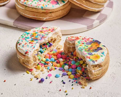An iced pinata sugar cookie broken in two with candy confetti and sprinkles spilling out, shown with three more cookies on a white and mauve checkered tray in the background.