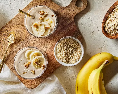  Top view of two tall glasses of banana oat tahini smoothie shown on a wooden cutting board with bowls of golden yellow sugar, sesame seeds, and dry oats with two unpeeled bananas.